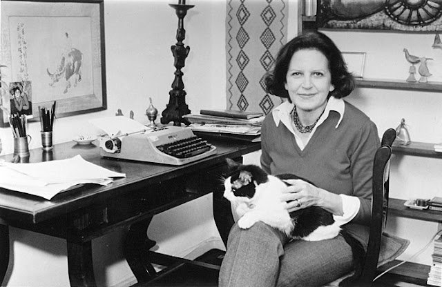Retrato em preto e branco de uma mulher sorridente com um gato bicolor no colo, sentada em seu escritório ao lado de uma mesa com uma máquina de escrever vintage.