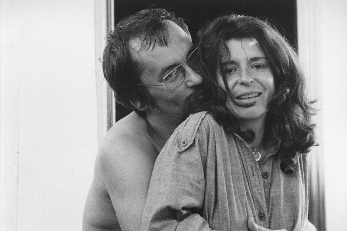 Casal em foto preto e branco, com o homem abraçando a mulher por trás e beijando seu pescoço enquanto ela sorri.