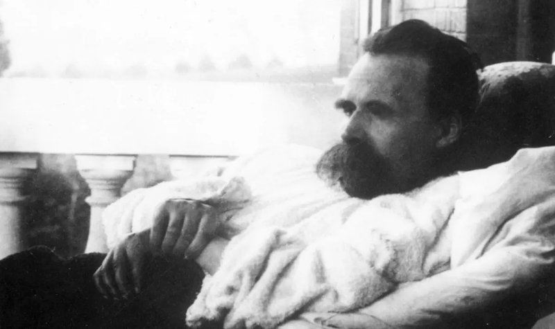 Retrato em preto e branco do filósofo Friedrich Nietzsche, com sua icônica barba e bigode, reclinado e olhando pensativamente para o lado. Ele está coberto por um cobertor, sugerindo um momento de descanso ou reflexão.
